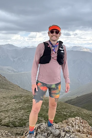 Ory Frazier standing on a rocky summit ridge in running kit, mountains behind