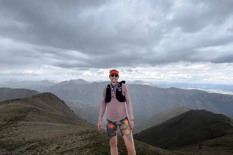 Ory Frazier on a high ridgeline in running kit, mountains and storm cloud behind
